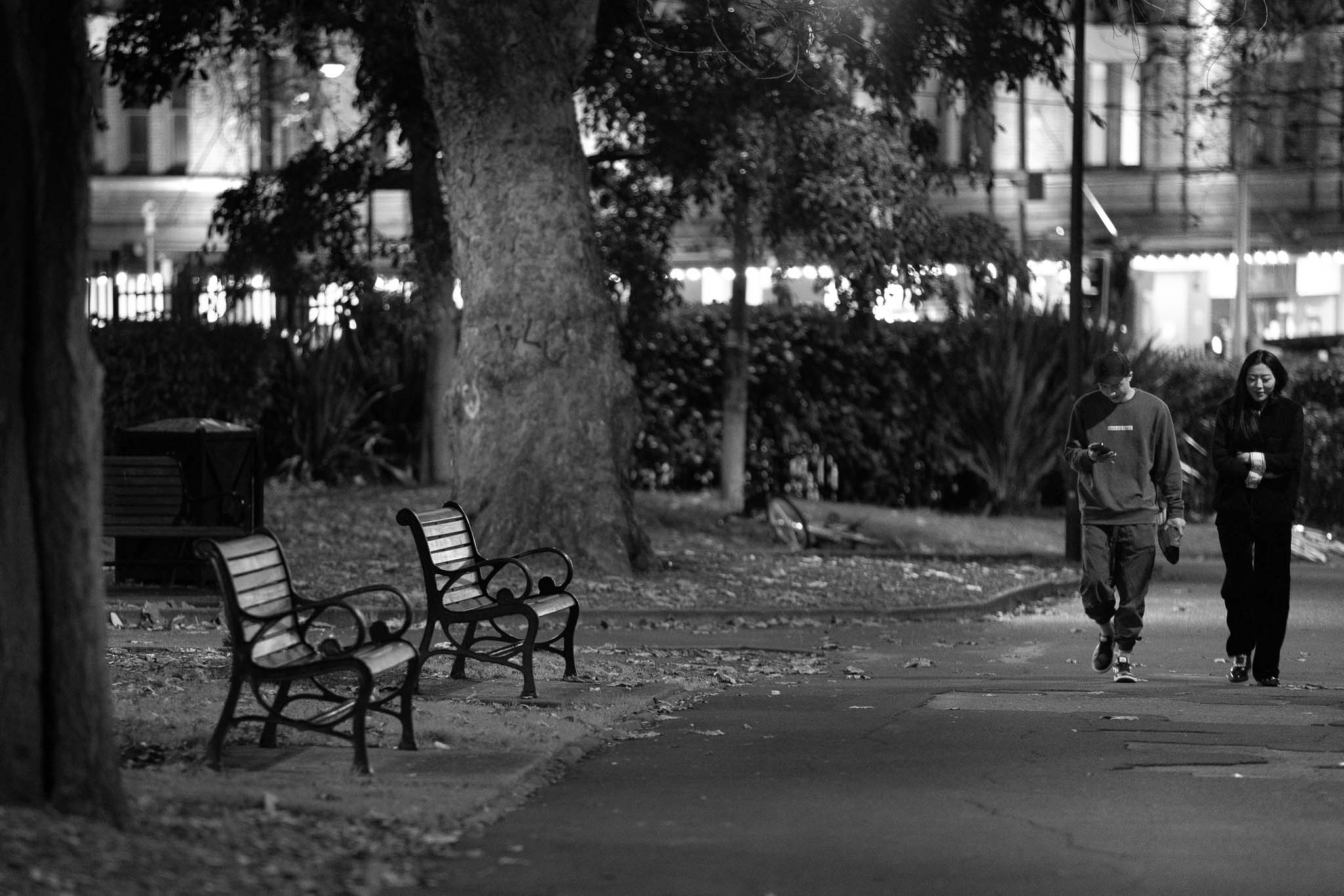 A couple mid-step walking up through Belmore Park at night, with Sydney's theatre district in the background. The man is looking down at his phone, while the woman is smiling with her arms crossed. Two empty park benches are to the side of the patchy path.