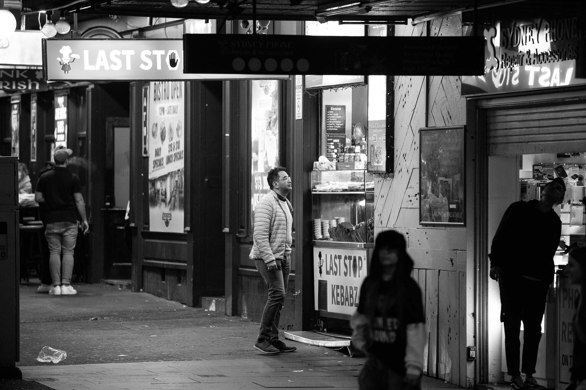 A guy in a puffer jacket and glasses is standing in a relaxed, limp pose out the front of Last Stop Kebabz, looking up at the menu. In the foreground, two people are in silhouette to the right, standing in front of a soon-to-close phone repair stall, while to the left, a group is visible out the front of an Irish pub, one man smoking.