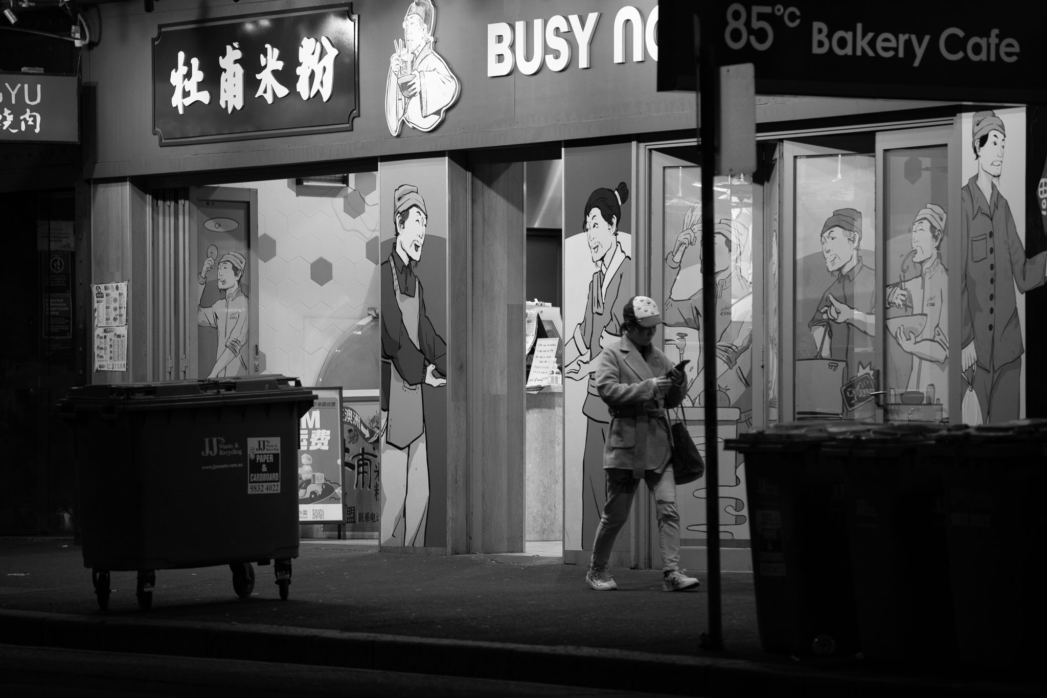 A woman is walking in front of a takeaway food store, wearing a cap, handbag, clad in a jacket, and looking down at her phone. The storefront is partially open, with the door panels and columns illustrated with drawings of cafe workers preparing food. Rubbish bins are visible in silhouette in the foreground.