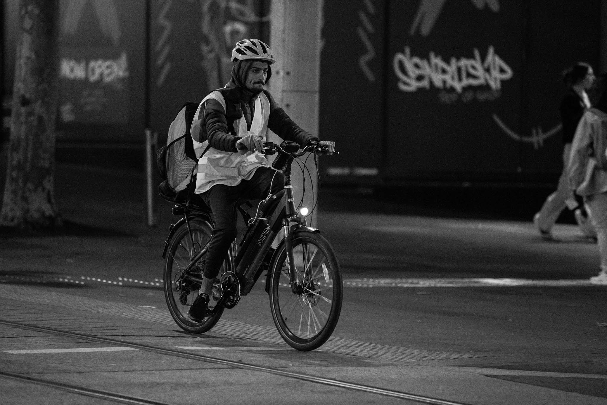 A tired, gruff-looking guy wearing a helmet, high-viz and a delivery company backpack is riding a bike along the street next to tram tracks. In the background, a couple of out-of-focus pedestrians can be seen wearing more relaxed clothing in front of a dark storefront.