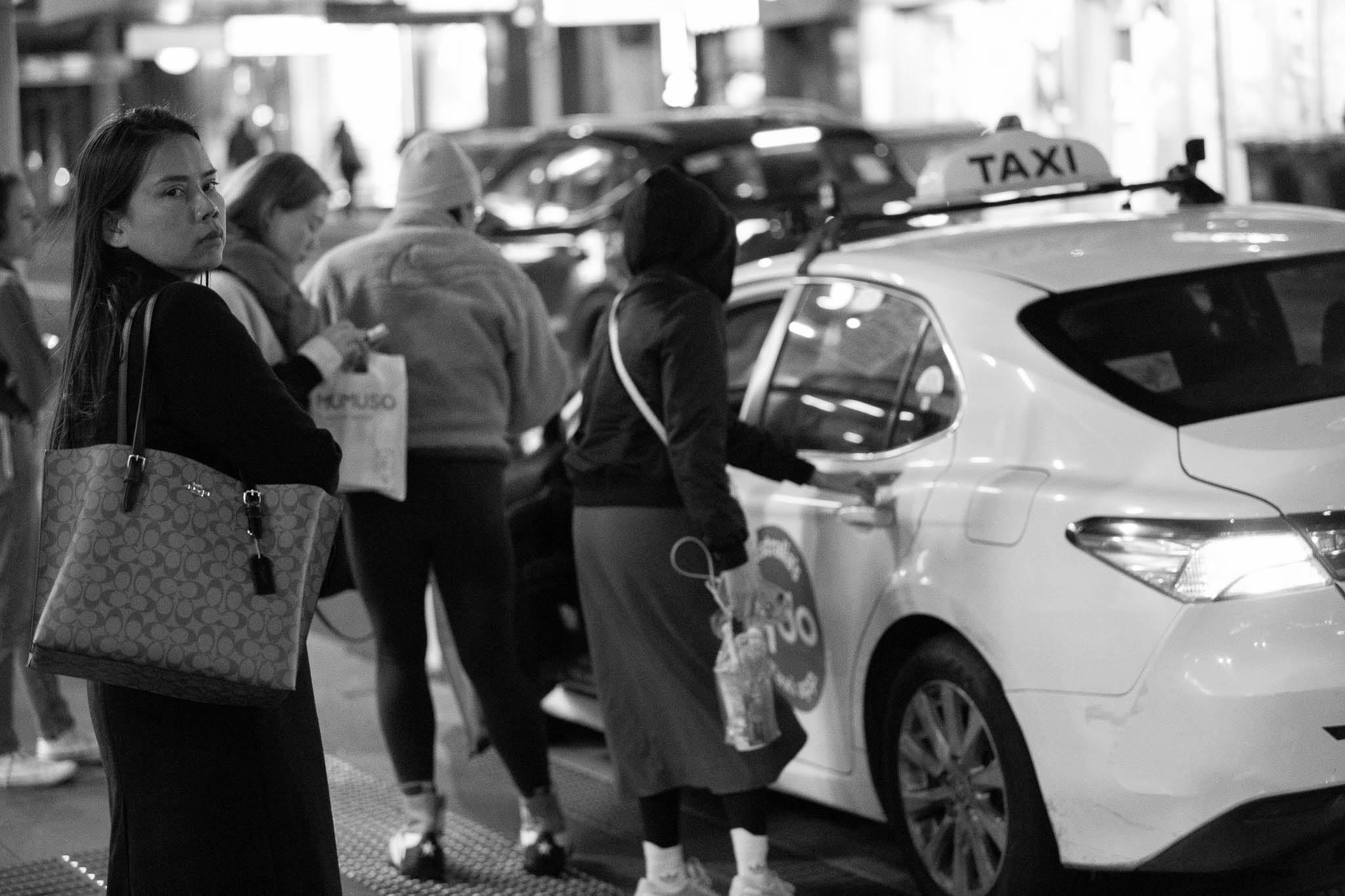 A crowd is queueing up to get into a taxi, with two actively getting into a cab, a third holding a shopping back and looking down at their phone, the trio out of focus in the background. A woman is visible to the left, in-focus, glancing back at the camera with a concerned look.