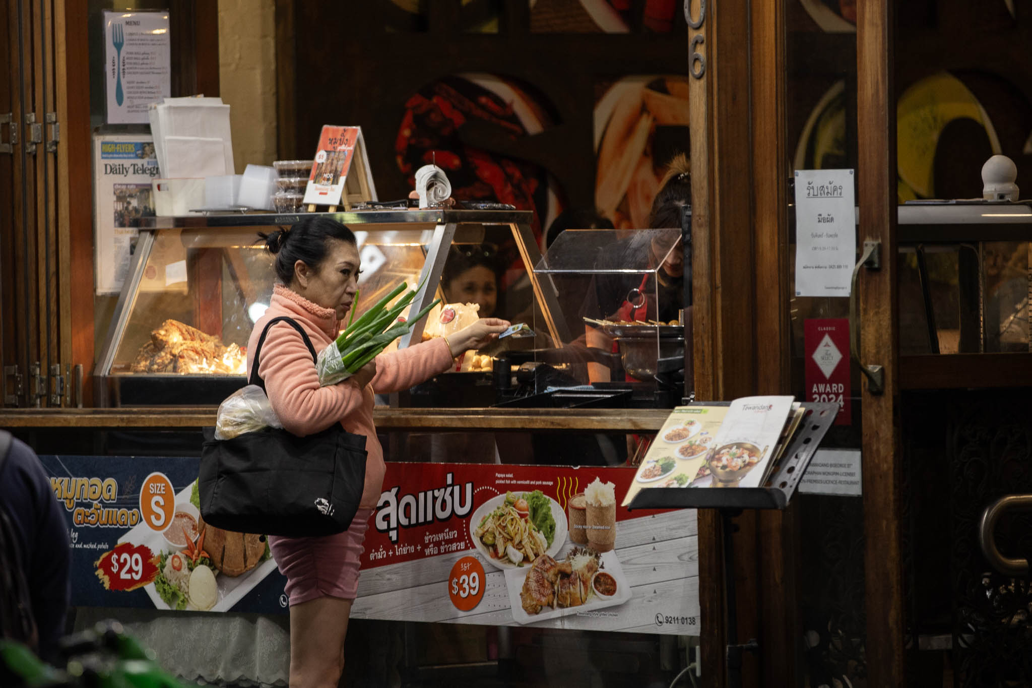 An older woman in a peach-coloured tracksuit has a black bag over her arm, a couple of long green veg under her arm, and is handing money to the cashier at a Thai restaurant's takeaway storefront. She is looking disapprovingly at something off-frame.