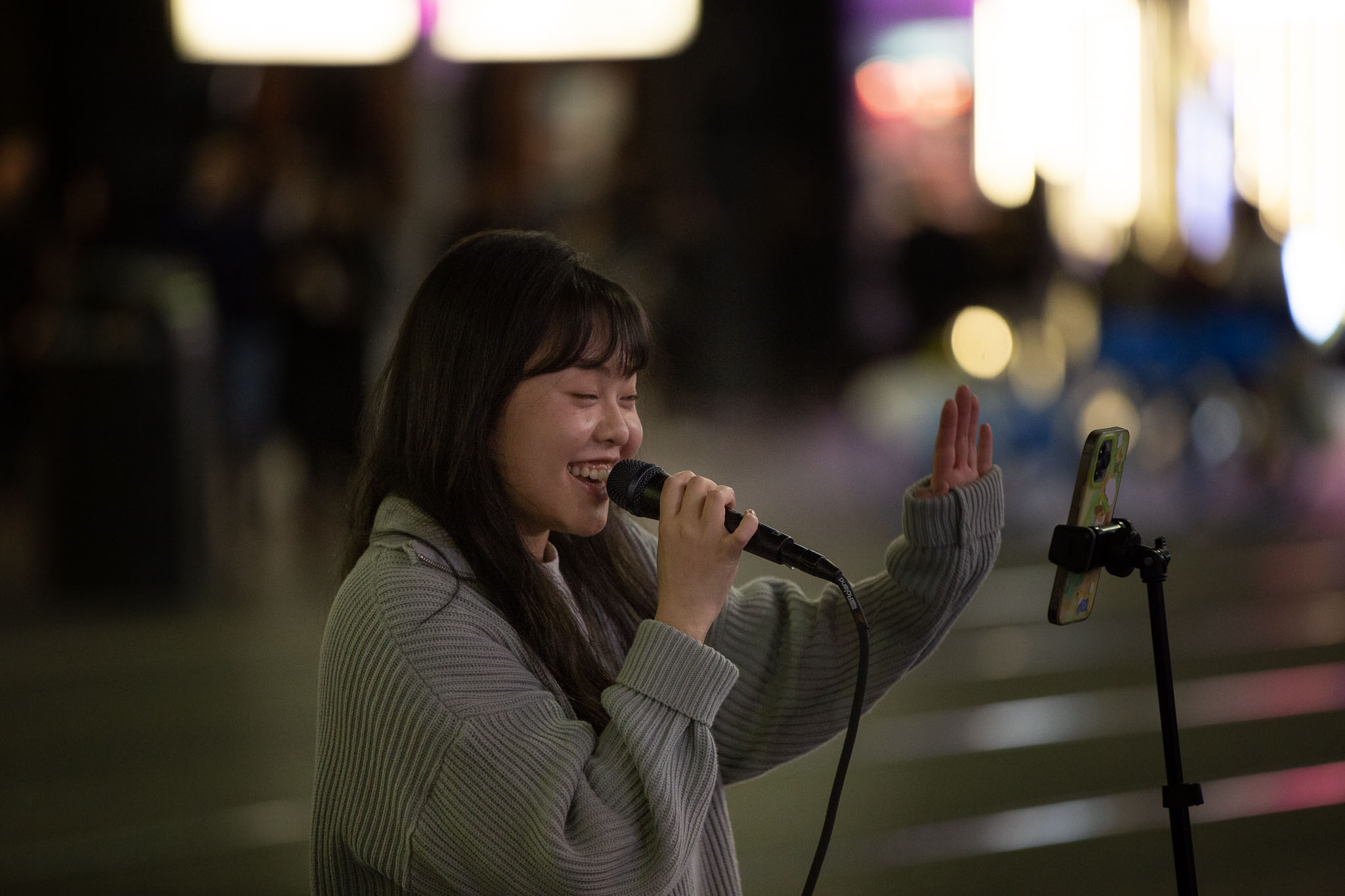 A woman is performing to a small crowd, with her phone on a stand showing the music. She has a wireless microphone to her lips, her hand up waving to someone, and a massive smile on her dial. The heavily-blurred lights of George St and the tramway are visible in the background.
