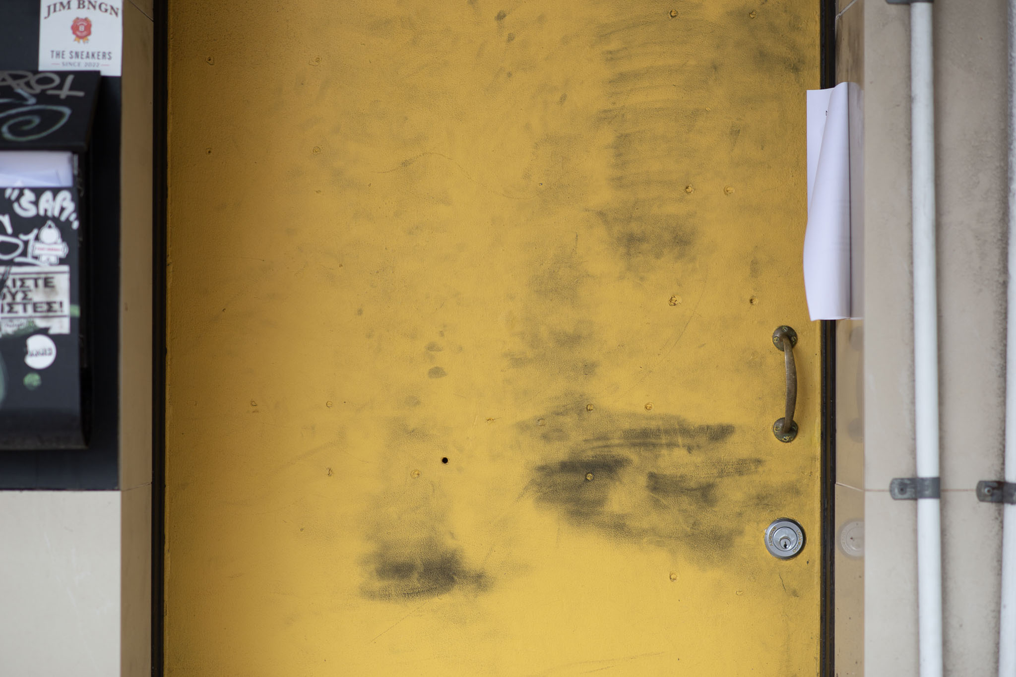 A bold yet plain, yellow door, covered with dark, grubby hand marks sits between a mailbox covered in graffiti and stickers, and PVC cable conduit. A folded-over printout of some announcement is taped to the wall on the right-hand side, and the door looks weary with age and use.