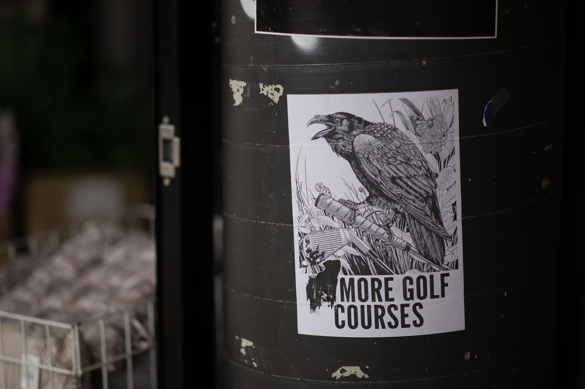 An A4 wheatpasted poster is stuck to a black-painted concrete column, white in colour with a loud, squawking crow perched atop the handle of a golf club. A second golfclub is upturned, blood covering the face of the club, with long foliage behind both clubs and the bird. A silhouette of the grim reaper appears alongside the title at bottom, 'More Golf Courses', written in block capitals. Behind the column and poster is an anonymous, out-of-focus basket of goods in front of a store.