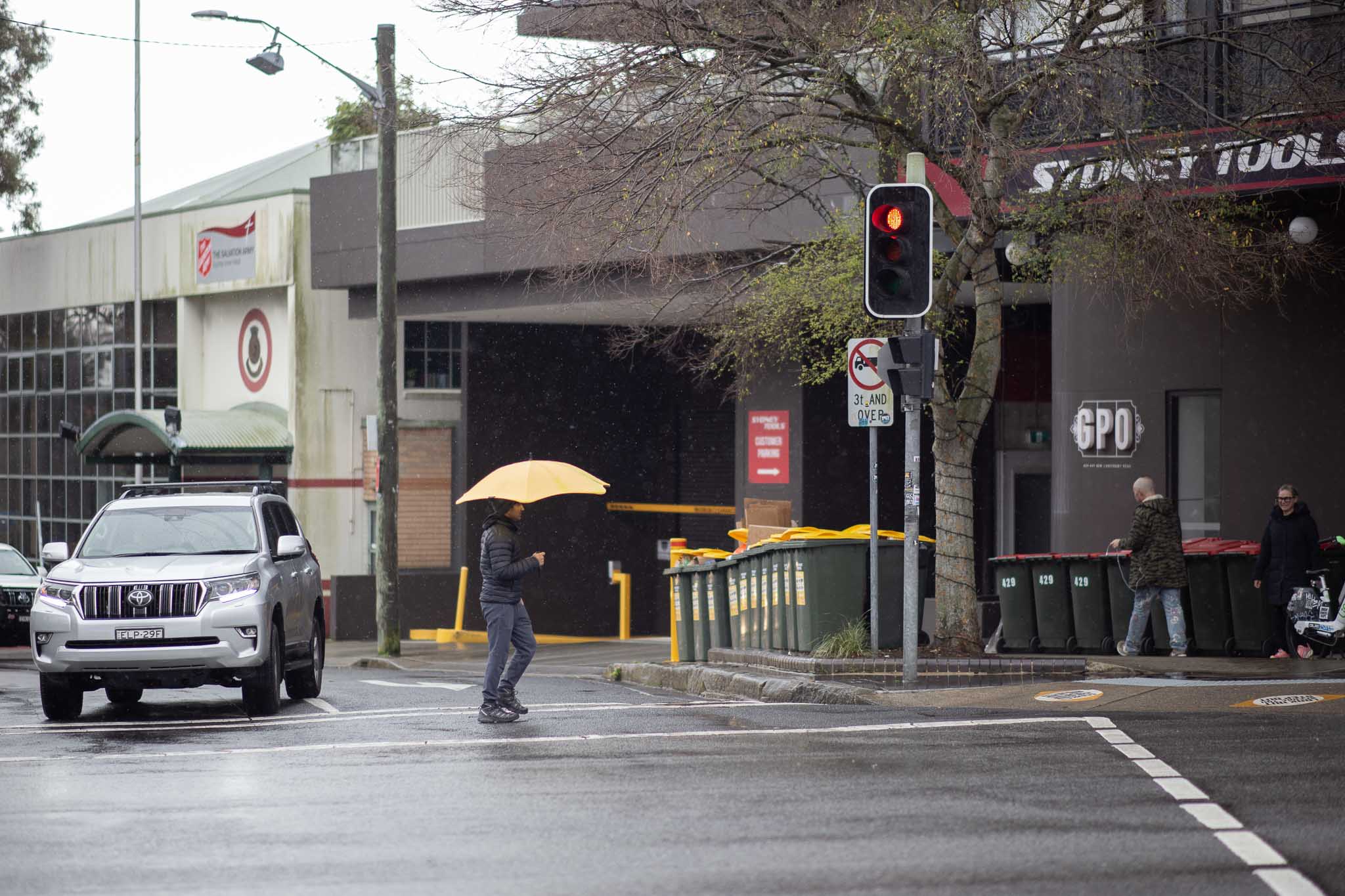 A street scene at a four-way intersection on an overcast, rainy day. A man in a puffy jacket is walking across a pedestrian crossing with a bright yellow umbrella, halfway across the road. To his side are a row of ten identical, regularly- and narrowly-spaced yellow-lidded garbage bins, green in colour.