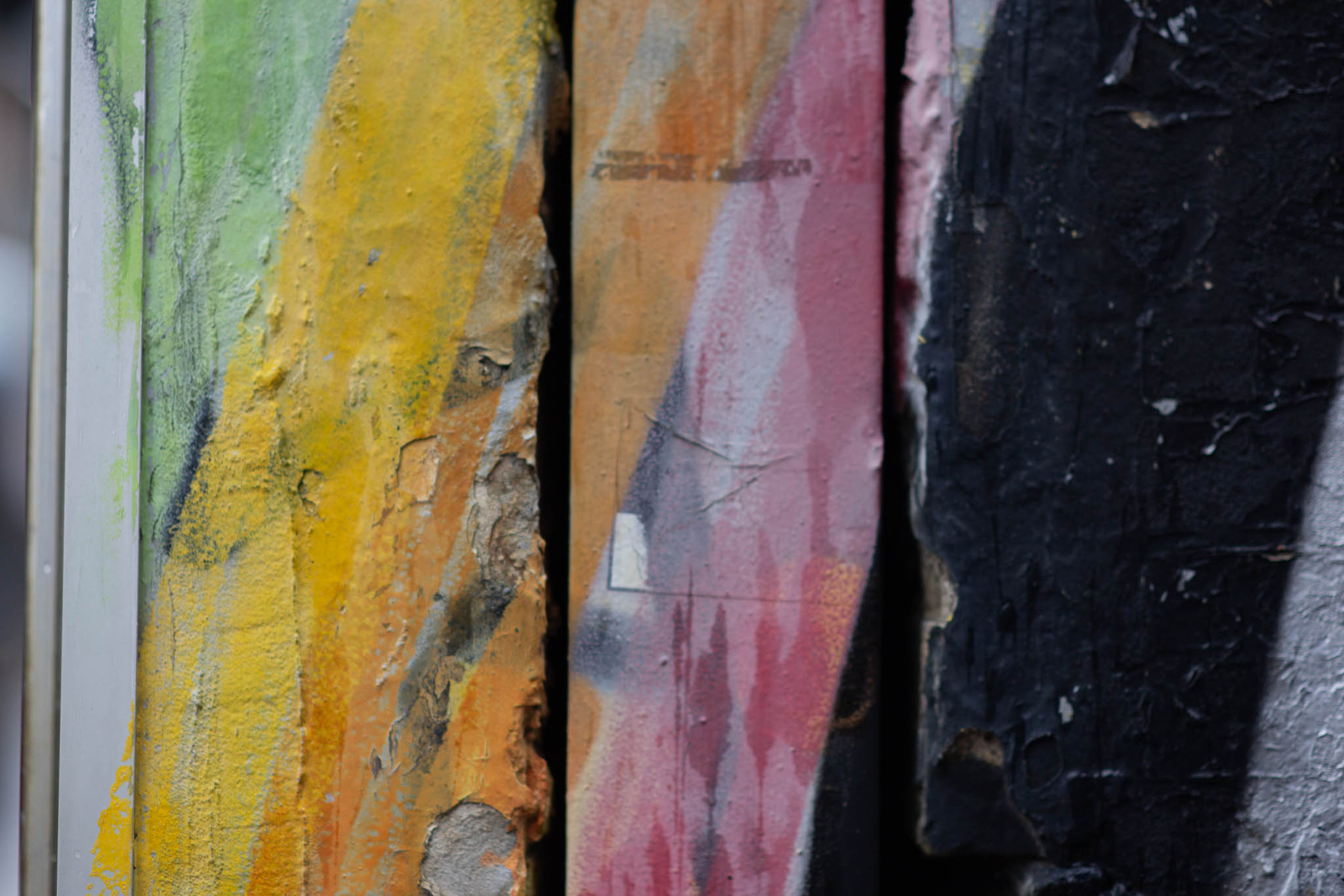 An abstract photo of a well-textured building wall in a laneway, the texture consisting of bricks covered with years and years of paint and muck. The wall is painted over with vivid swaths of line green, yellow, orange, white, pink, black and silver paint, in mostly-vertical, slightly-diagonal patches. 