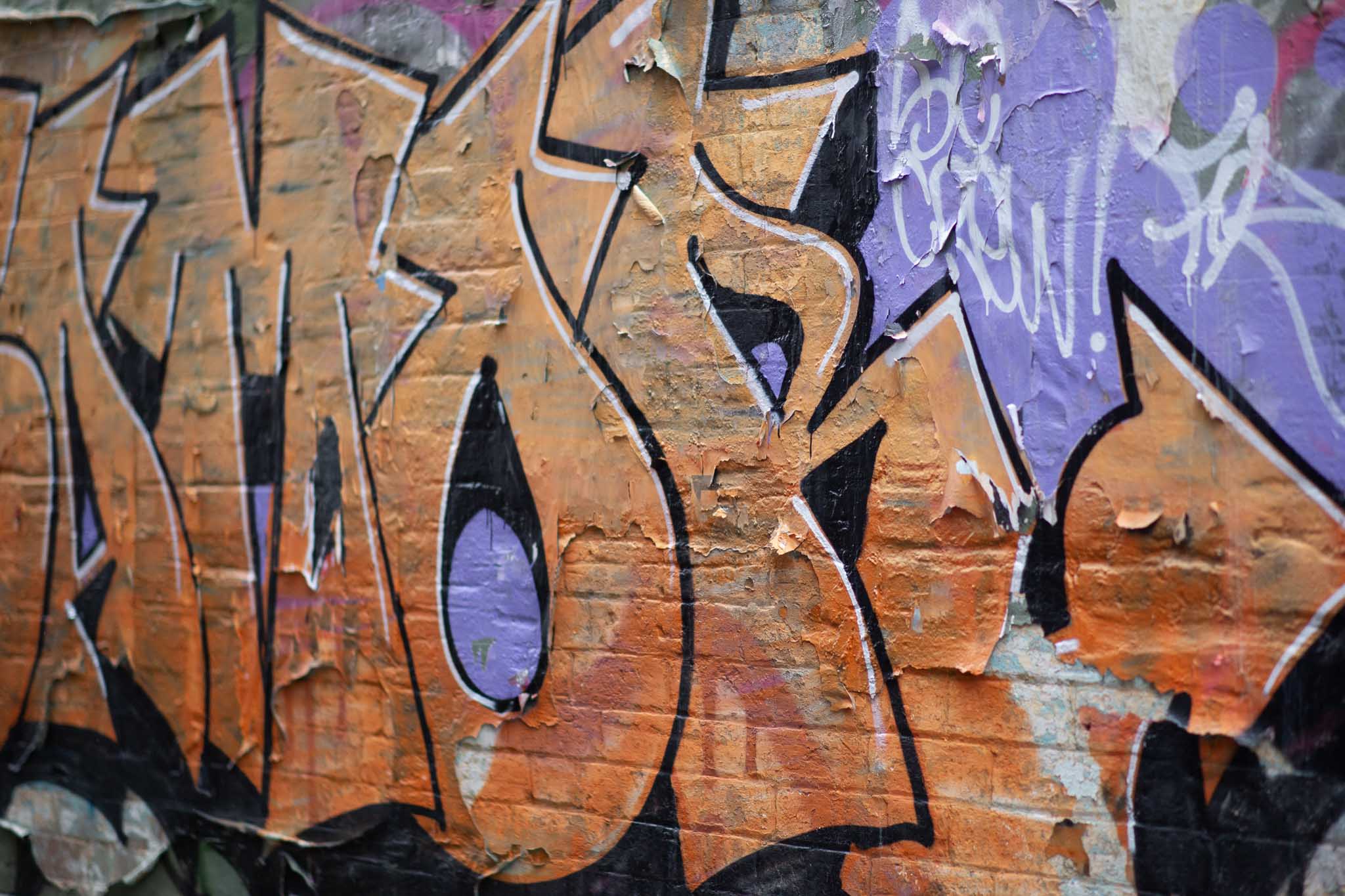 A photo of vividly-coloured, large tagging on a brick wall in a laneway. The text is made of large, orange block letters with sharp angles and organic curves, outlined with a white highlight to the upper right of the blocks, and framed in a black outline and drop shadow to the lower of the text. The background is a desaturated purple that contrasts with the warmth of the orange.