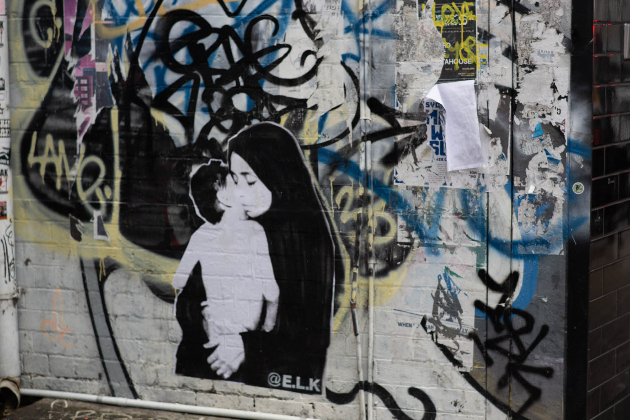 A stencil-style graffiti poster is stuck to the wall, showing a woman in a dark-coloured hijab cradling her child, the child's face hidden against her body. Behind the stencil, various tags are on the brick-textured wall with a white background, yellow, blue and black tags covering half the wall. The stencil has '@E.L.K.' written in the corner.