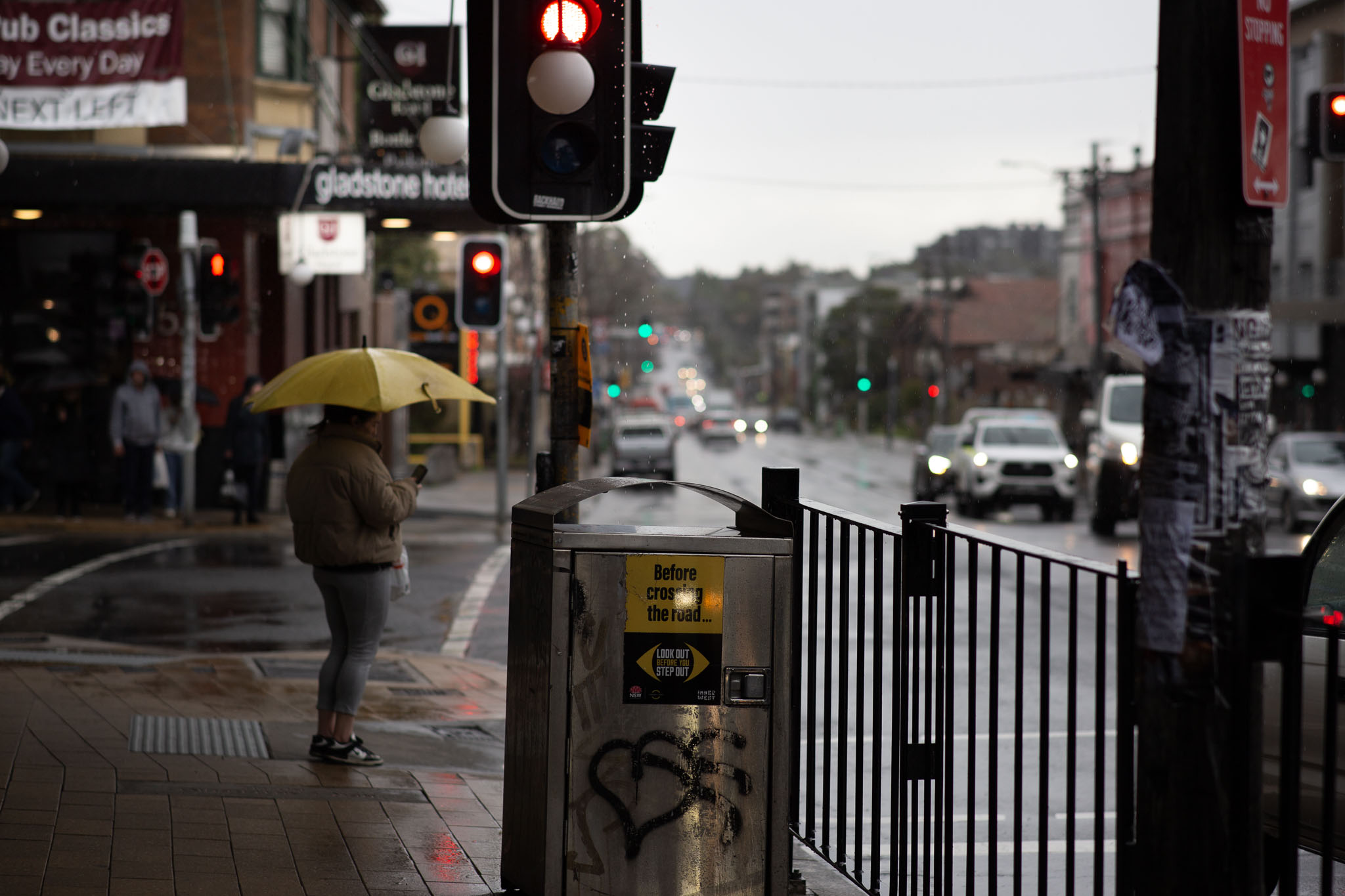A person in a puffy rain jacket and yoga pants is waiting at a pedestrian crossing, holding a bright yellow umbrella. Behind them, a couple of people wait across the road to cross the other direction. In the foreground is a metal rubbish bin with a sticker reading 'Before crossing the road..'