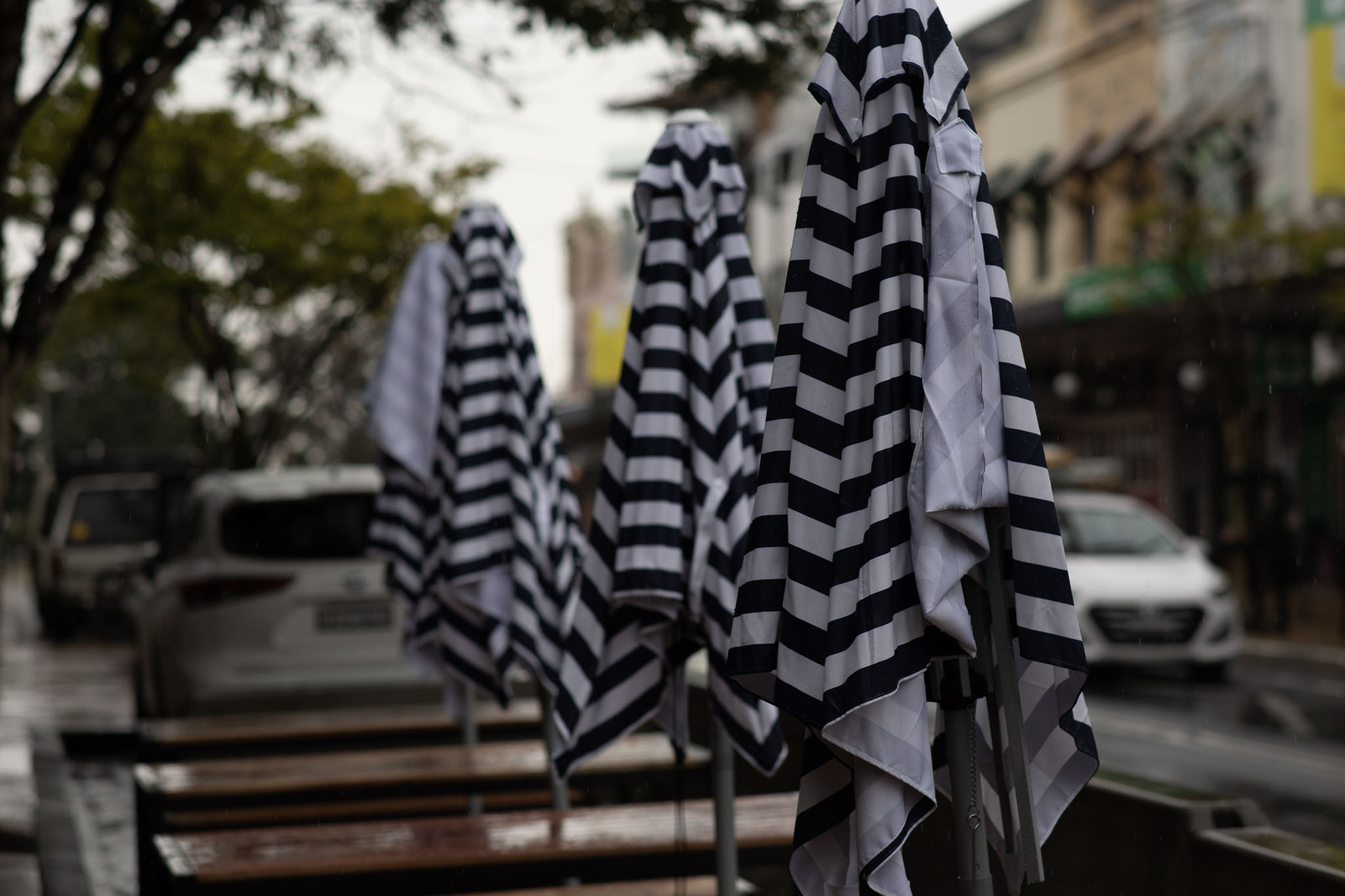 Black-and-white striped umbrellas are folded up, sitting atop a bunch of streetside tables outside a cafe. The photo shows a street scene with a number of parked cars behind the streetside tables, on what is a clearly wet and overcast day, the black-and-white umbrellas looking like a folded-up keffiyeh in black-and-white, as adopted by Palestinians.